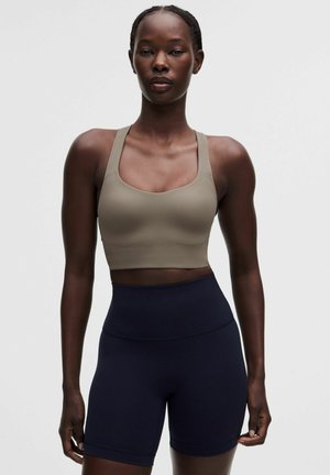 Donna che indossa un reggiseno sportivo taupe e pantaloncini sportivi blu navy a vita alta, in piedi su uno sfondo bianco semplice.