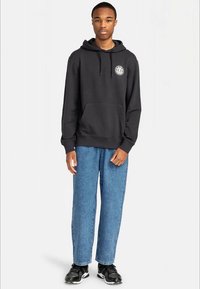 Sweatshirt preto de algodão com um bolso frontal e detalhe de logotipo, combinado com calças de ganga azul claras e tênis atléticos pretos.