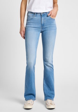 Kobieta w jasnoniebieskich bootcut jeansach, białych trampkach i białym włożonym do spodni t-shircie, z jedną ręką w kieszeni.
