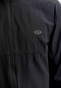 Veste noire avec fermeture éclair à l'avant et petit logo réfléchissant sur la poitrine gauche, en tissu texturé.