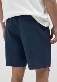 Pantaloni corti blu navy con un tessuto testurizzato, caratterizzati da una tasca posteriore con due bottoni, taglio dritto e una vita che si adagia comodamente.