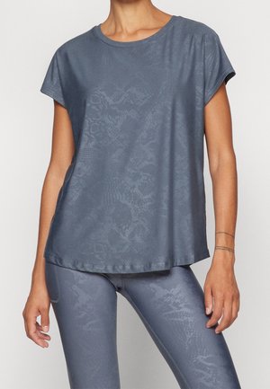 Kvinde iført en løs mørkegrå kortærmet skjorte og matchende leggings med et diskret slangehudsmønster på en ensfarvet baggrund.