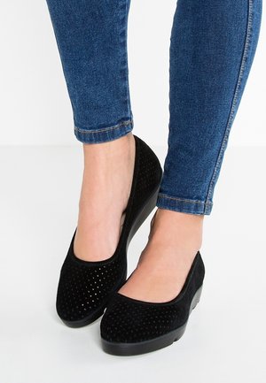 Czarne perforowane buty wsuwane z okrągłym noskiem, teksturowaną cholewką i niskim obcasem w kształcie klinu, zestawione z niebieskimi dżinsami.