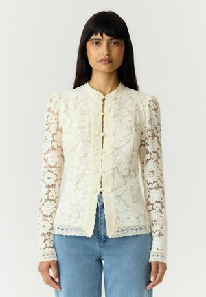 Femme aux longs cheveux foncés portant un chemisier en dentelle crème à manches longues avec des boutons décoratifs et un jean bleu, debout devant un fond uni.