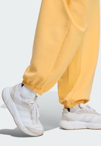 Nohy v bielych teniskách a bielych ponožkách, s voľnými žltými nohavicami zasiahnutými v členkoch na svetlom pozadí.