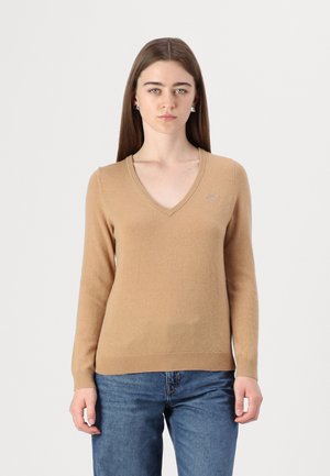 Beige V-Ausschnitt Pullover mit langen Ärmeln, aus weichem Material. Verfügt über ein kleines Logo auf der Brust und eine figurbetonte Silhouette, kombiniert mit blauen Jeans.