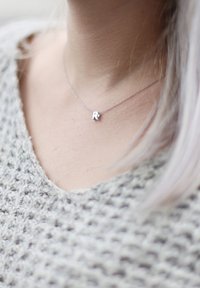 Zilveren hanger ketting met een "R" charm, op een dunne ketting. Draagt boven een gestructureerde lichtgrijze gebreide trui met een V-hals ontwerp.