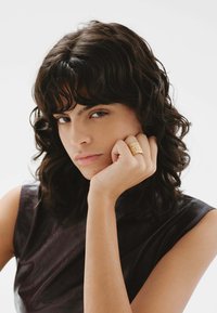 Anillo texturizado dorado llevado en la mano, con una blusa sin mangas oscura visible. La modelo tiene el cabello rizado oscuro y una expresión neutra.