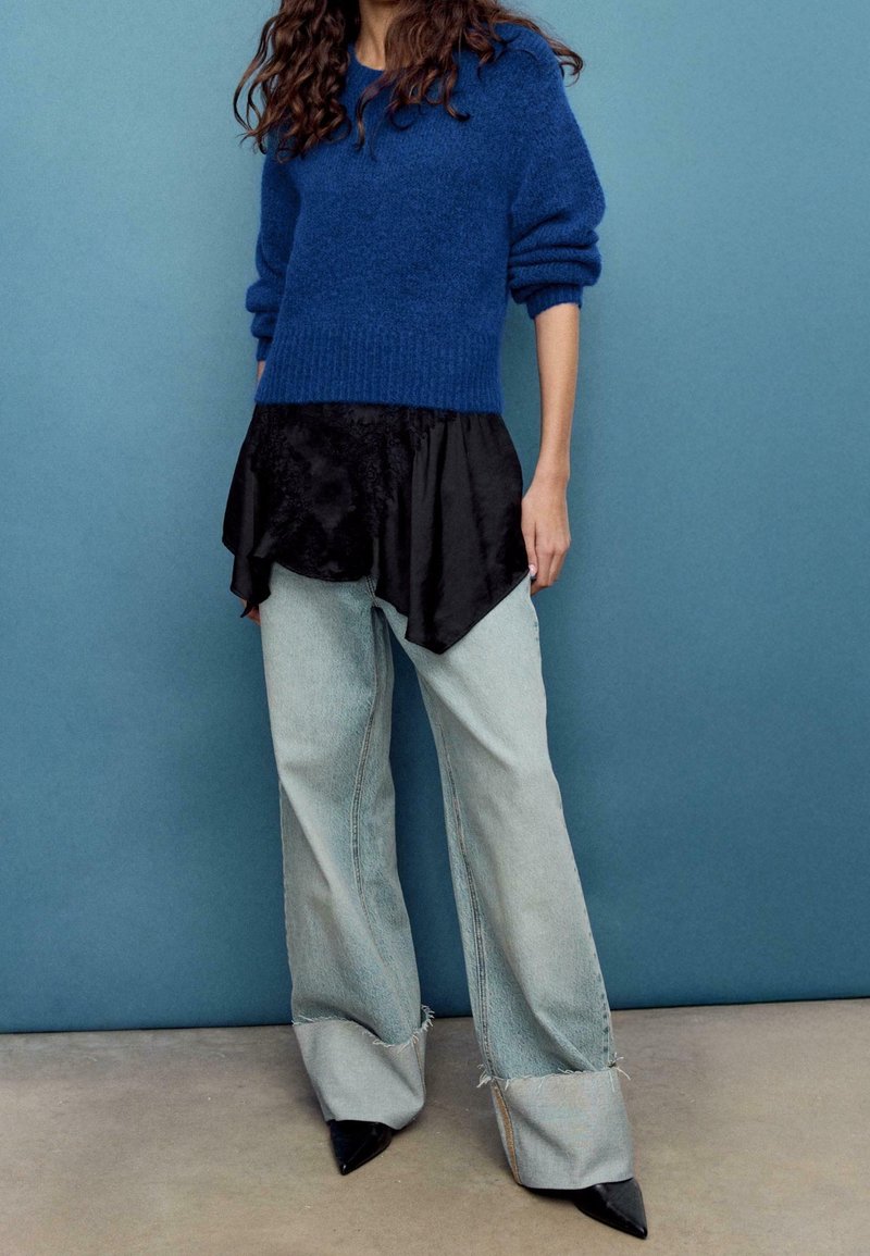 Femme portant un pull bleu, un top en soie noire en plusieurs couches, un jean denim clair à jambes larges avec des ourlets bruts, et des chaussures noires à bout pointu devant un mur bleu.