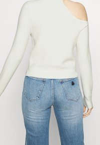 Light green ribbed sweater with one shoulder exposed, paired with light blue denim jeans featuring a triangular accent on the back pocket.