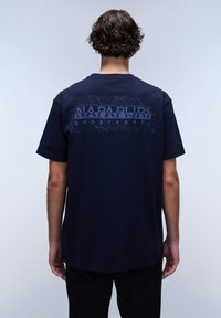 Tmavě modré bavlněné tričko s texturovaným grafickým potiskem na zádech s nápisy "NAPAPIJRI" a "geographic" v kontrastních barvách. Krátké rukávy.