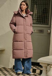 Palton lung, tip puffer, într-o nuanță moale de mov, având un guler înalt, nasturi tip snap și secțiuni mari quilted. Asortat cu blugi cu picior larg și încălțăminte ușoară.