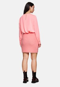 Rosa Bluse mit einem Schlüssellochdetail am Rücken, kombiniert mit einem rosa strukturierten Minirock. Das Model trägt knielange schwarze Socken und braune Stiefeletten.