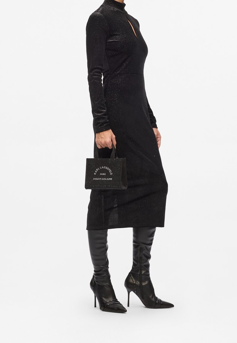 Femme portant une robe noire à manches longues et des bottes noires à talons hauts, tenant un petit sac à main noir Karl Lagerfeld.
