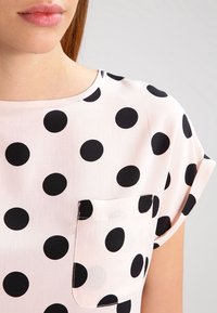 Ljusrosa kortärmad blus med svarta prickar, med en ficka på vänster sida. Slätt tyg med en subtil lyster.