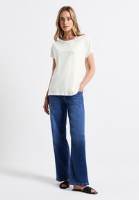 Camiseta de manga corta en color crema claro que presenta un estampado texturizado. Combinada con jeans de pierna ancha azules y sandalias negras de tipo slip-on. Diseño minimalista.