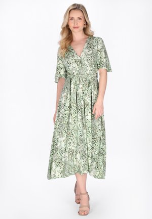 Nő hosszú szőke hajjal, aki zöld és bézs mintás, rövid ujjú midi ruhát visel, valamint bézs színű, magas sarkú szandált.