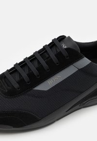 Chaussures de sport noires en mélange de daim et de mesh, avec un bout rond, des lacets noirs et un détail logo métallique.