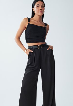 Schwarzes, kurzes Oberteil mit breiten Trägern und metallischen Akzenten, kombiniert mit hoch taillierten, weiten schwarzen Hosen mit einem Gürtel und Falten.