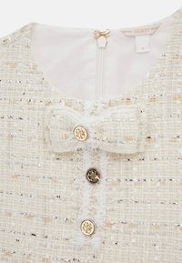 Weiße Tweed-Bluse mit Schleifendetail, goldenen Akzenten und sichtbarem Reißverschluss. Goldene und silberne Fadenmuster verzieren die Textur.