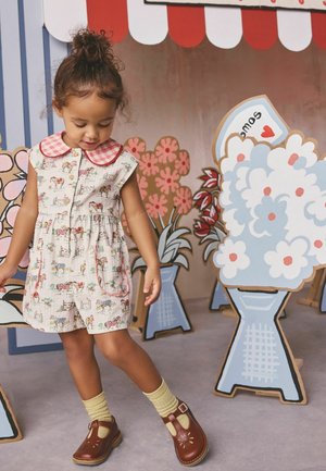 Dziewczynka w romperze w wzory i brązowych butach stoi obok kolorowych kartonowych kwiatów pod czerwono-białym pasiastym markizem.