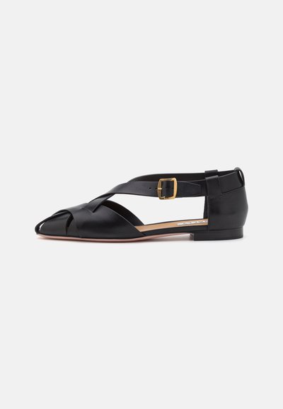 Scarpa piatta in pelle nera con punta aperta, cinturini incrociati e cinturino alla caviglia regolabile con fibbia dorata; finitura liscia.