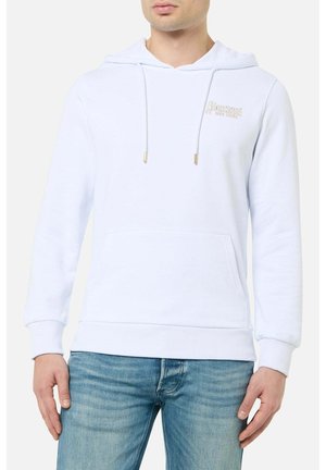 Mann trägt schlichtes weißes Hoodie mit Fronttasche und hellblauen Jeans, steht vor einfarbigem Hintergrund.