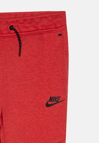 Sarkanas sporta bikses, kas izgatavotas no struktūrēta auduma, ar melnu Nike logo kreisajā apakšējā stūrī un aukliņu viduklī.