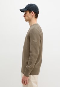 Homme en pull beige texturé et casquette noire, vu de profil sur un fond blanc uni.