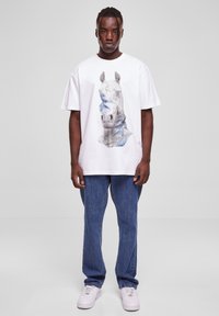 Baltas T-krekls ar stilizētu zilu un pelēku zirga grafiku, apvienots ar ziliem džinsiem un baltiem sporta apaviem. Brīva piegriezuma un kokvilnas materiāls.