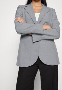 Šedý a černý kostkovaný blazer s výstřihem do písmene V, přizpůsobeným střihem a zakřiveným spodním lemem, nošený přes bílé tričko, v kombinaci s černými kalhotami.
