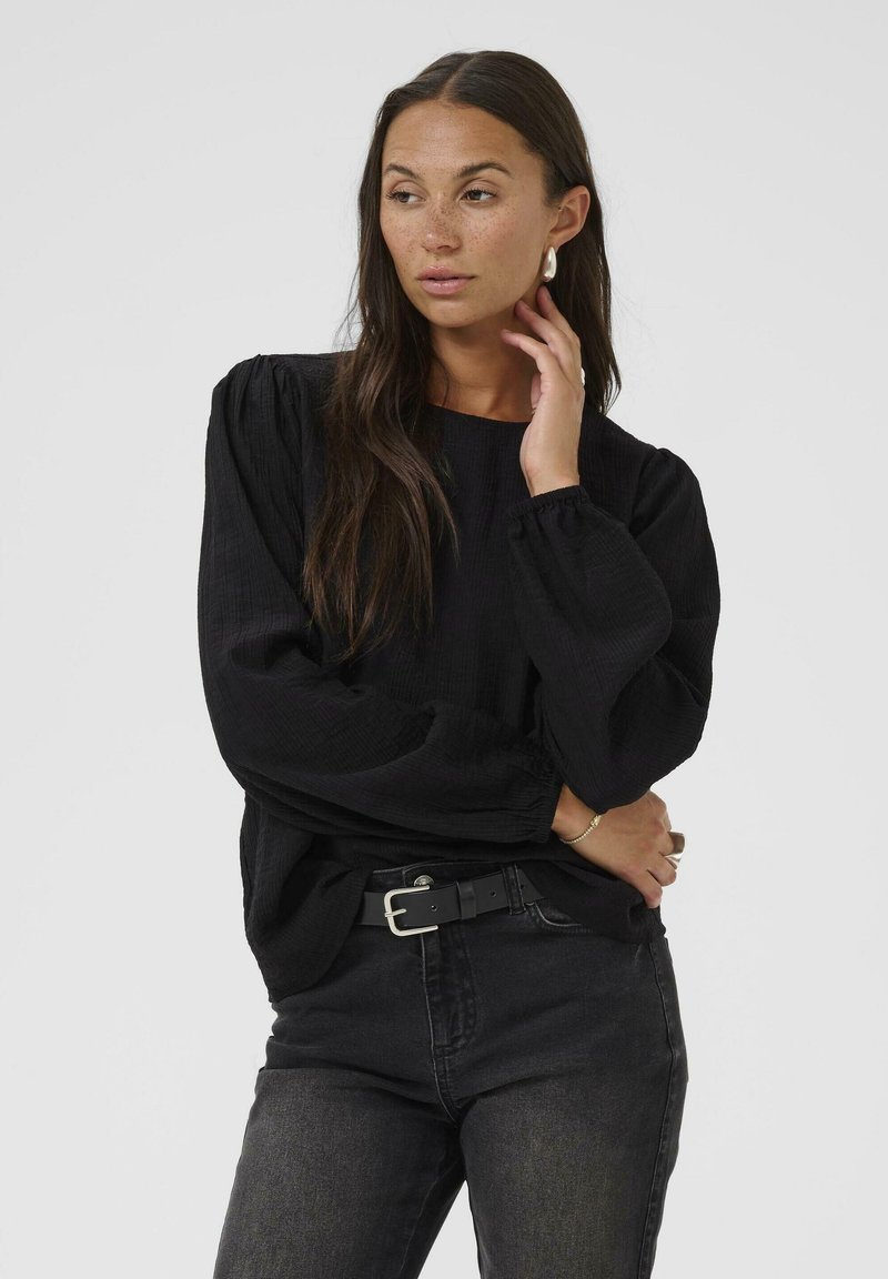 Blouse noire à manches longues en tissu texturé, coupe décontractée et poignets élastiqués, assortie à un jean foncé et une ceinture noire.