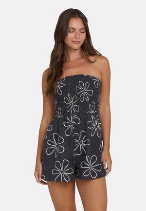 Donna con capelli castani lunghi che indossa una tuta nera senza spalline con motivi floreali bianchi, in piedi davanti a uno sfondo chiaro e semplice.