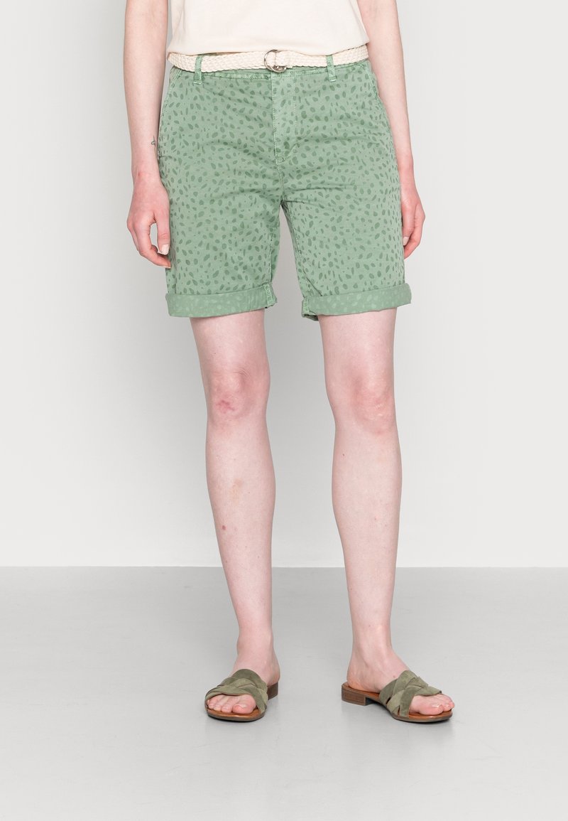 Pantalones cortos de algodón verde con un sutil patrón de puntos, puños enrollados y bolsillos frontales. Combinados con sandalias de tiras color oliva.
