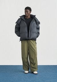 Νέος άνδρας με μαύρο μπουφάν puffer, μαύρο πουκάμισο, φαρδιά παντελόνια ελιάς και λευκά αθλητικά παπούτσια, στέκεται πάνω σε μπλε χαλί.