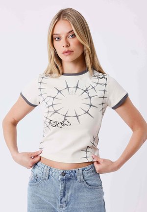Jeune femme aux longs cheveux blonds portant un t-shirt blanc avec un motif circulaire gris épineux et un jean bleu, debout sur un fond blanc.