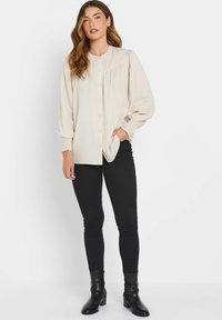 Blouse beige à boutons avec des manches longues et un design froncé à l'avant. Associée à un jean skinny noir et des bottines, créant un look élégant.
