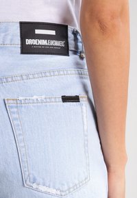 Des jeans en denim bleu clair présentent une étiquette tissée noire avec "DRDENIM JEANMAKERS" à l'arrière. Le tissu a une texture lisse et un léger dégradé.