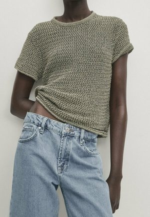 Personne portant un haut tricoté beige texturé à manches courtes et un jean taille haute bleu clair, debout avec une main dans le dos.