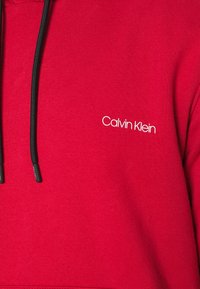 Sudadera roja hecha de tela suave, con un cordón negro y un pequeño logo blanco "Calvin Klein" en el pecho.