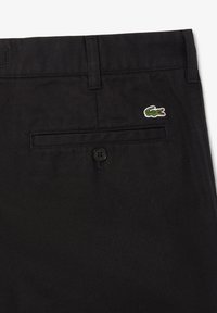 Pantaloni in cotone neri con una texture liscia, dotati di una tasca posteriore con bottone e di un piccolo logo di coccodrillo verde ricamato.