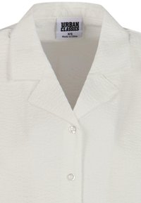 Hvit kortermet skjorte med teksturert stoff, nothals og en rekke med tre knapper. Merket leser "Urban Classics," størrelse XS.