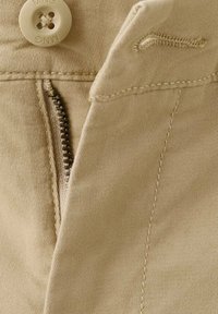 Prim-plan cu pantaloni bej, cu fermoar parțial deschis, nasture cu gravură "MNG" și petic cusut pentru nasture pe o bandă de talie din material textil.