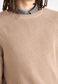 Beżowy sweter dziany o prążkowanej fakturze i okrągłym dekolcie, nałożony na szary podkoszulek z kołnierzykiem, z raglanowymi rękawami.