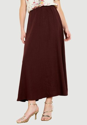 Žena má na sebe dlhú tmavohnedú sukňu v kombinácii so zlatými sandálmi na vysokom podpätku s remienkami, stojí pred jednofarebným pozadím.