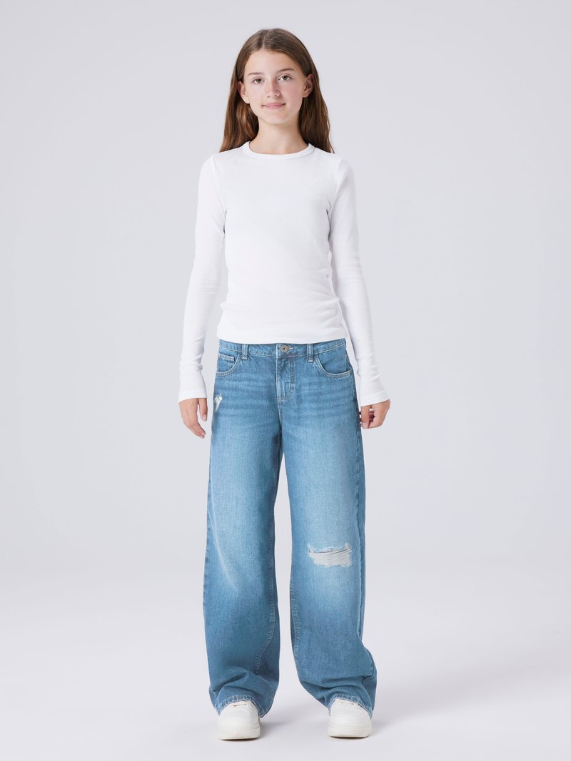 Tricou alb cu mânecă lungă și model rigat, asociat cu blugi largi decolorati, de un albastru deschis, și adidași albi. Ținută simplă și lejeră, cu texturi de bază.