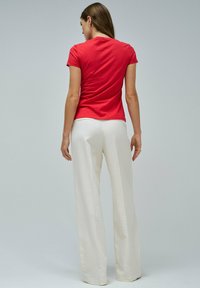 Mujer de espaldas con una camiseta corta roja y pantalones anchos blancos, contra un fondo neutro liso.