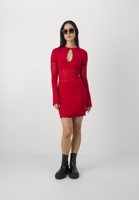 Donna che indossa un mini abito rosso a maniche lunghe con dettagli in pizzo e scollatura a chiave, stivali neri chunky e occhiali scuri, in piedi contro uno sfondo semplice.