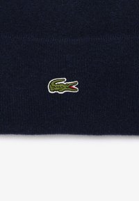 Bonnet tricoté marine en tissu doux, arborant un petit logo de crocodile vert brodé avec des accents rouges sur le devant.