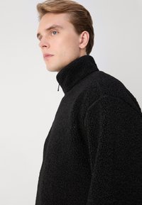 Zwarte fleece pullover met hoge kraag, gestructureerd oppervlak en ritssluiting. Heeft een ontspannen pasvorm en ingesette mouwen voor comfort.